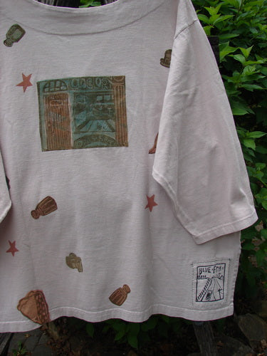1993 Resort PJ Top Diner Treat Tea Dye Size 2, featuring an abstract storefront print, brown icons, red stars, and a BLUE FINE graphic, hangs against lush foliage.