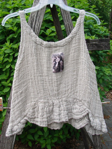 Magnolia Pearl Fisherman's Weave Ribbon Neck Top Flaxen OSFA, featuring crinkled, gauzy fabric with ruffled hem, displayed on a wooden ladder against leafy shrubs, includes a black-and-white portrait appliqué on the front.