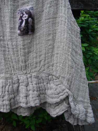Magnolia Pearl Fisherman's Weave Ribbon Neck Top Flaxen OSFA, hand-knit with gathered ruffled hem, displayed on wood with a decorative tag featuring an illustrated face, set against outdoor foliage.