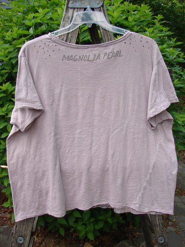 Magnolia Pearl Cotton Jersey Boyfriend Tee Queen of Hearts Pink Dust OSFA, featuring stenciled back text, shoulder distressing, and flutter edges, displayed on a wooden ladder amidst garden foliage.