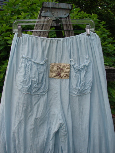 Magnolia Pearl Silk Voile Window Lace Ruffle Pocket Bloomer Sky OSFA displayed on a hanger, featuring gathered patch pockets, a beige insect motif patch, and intricate silken ribbon tie bottoms against a green foliage backdrop.