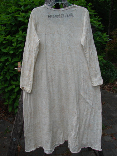Magnolia Pearl Cotton Dylan Dress Softest Floral Naranja OSFA, a floor-length cream bohemian duster with faint paisley print, long sleeves, and distressed details, hanging outdoors on a clear plastic hanger.