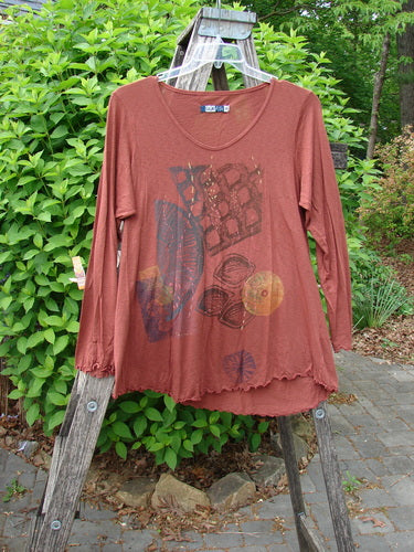 Barclay Modal Long Sleeved A Lined Tee Fall Woods Cassia Size 0, draped over a wooden ladder, showcasing its rust-brown hue, graphic front, wide neckline, and long sleeves against green shrubbery.