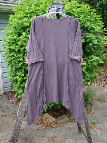 Barclay Wildflower Tunic Floral Sprig Fig Size 0: A drapey top with raw-edged front panel, dippy sides, flutter hemline, scooped neckline, and exterior drop pockets, displayed on a wooden stand outdoors.