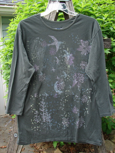 Barclay Long Sleeved Studio Tee Moon Gull Garden Ash Black Size 0 displayed on a wooden hanger, featuring a whimsical bird motif with scattered flowers against a leafy shrub background, showcasing artistic craftsmanship.
