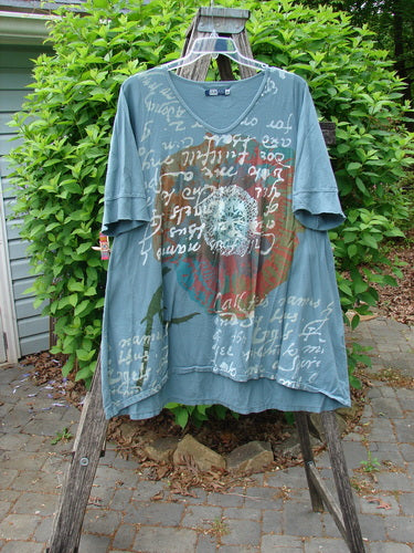 Barclay Three Quarter Sleeved Happiness Tunic Floral Notes Dusty Sky Size 0 displayed outdoors, featuring cursive white lettering, faded red heart motif, bird silhouettes, rounded V-shaped neckline, and drawcord back.