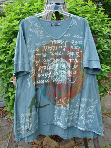 Barclay Three Quarter Sleeved Happiness Tunic Floral Notes Dusty Sky Size 0, showcasing a loose-fit design with elbow-length sleeves, central circular motif, and unique hemline, hanging outdoors.