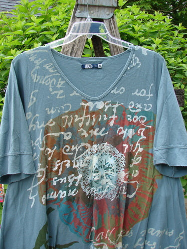 Barclay Three Quarter Sleeved Happiness Tunic Floral Notes Dusty Sky Size 0 displayed on a wooden trellis, showcasing its circular mandala design, hand-script text, and unique hemline cut.