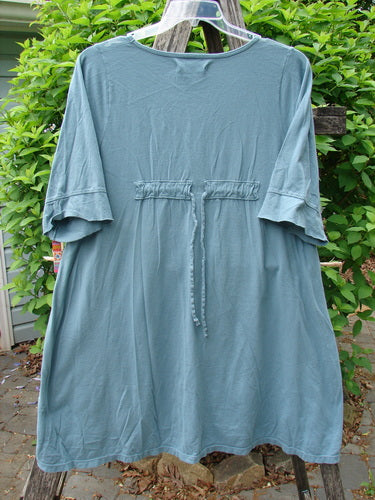 Barclay Three Quarter Sleeved Happiness Tunic Floral Notes Dusty Sky Size 0 features a loose fit with elbow-length sleeves, a drawstring back, and a high-low hemline, draped outdoors on a wooden frame.