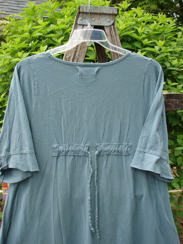 Barclay Three Quarter Sleeved Happiness Tunic Floral Notes Dusty Sky Size 0, featuring a drawstring back and rounded neckline, displayed on a hanger against a leafy trellis backdrop.