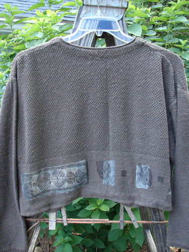 1994 Crop Cardigan Sweater Topiary Humus OSFA: The image shows a cropped cardigan sweater with longer sleeves, a V-shaped neckline, stoneware buttons, and intricate knit textures, hanging outdoors on a swing.