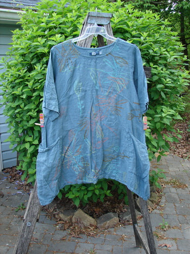 Barclay Linen Side Pocket Urchin Dress Mixed Tulip Garden Dusty Sky Size 0 hangs on a wooden ladder, showcasing its short sleeves, rounded neckline, exterior drop pockets, and a swaying lower hemline.