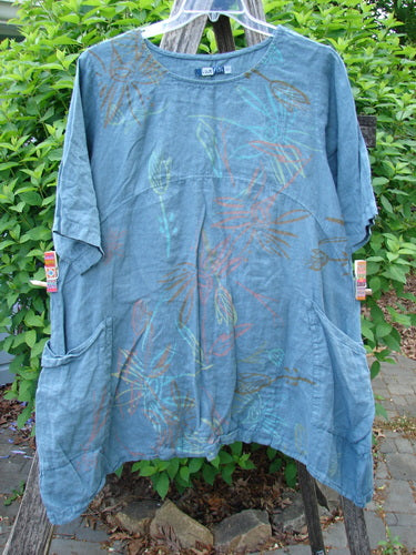 Barclay Linen Side Pocket Urchin Dress in Dusty Sky hangs in a garden, showcasing its round neckline, fern-print fabric, empire waist seam, and side pockets, embodying Bluefishfinder's vintage, creative style.