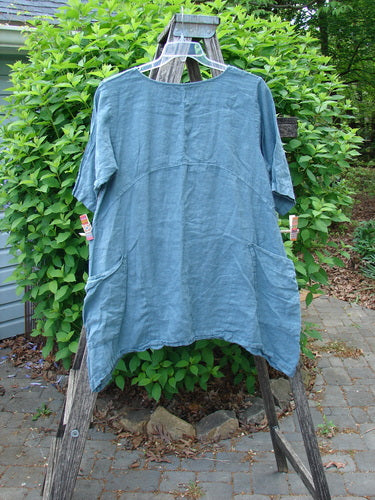 Barclay Linen Side Pocket Urchin Dress in Dusty Sky, showcasing mixed tulip garden design, hangs on a ladder; features include front patch pockets, rounded neckline, and empire waist seam. Size 0.
