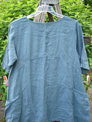 Barclay Linen Side Pocket Urchin Dress in Dusty Sky hangs on a ladder, featuring short sleeves, empire waist, exterior drop pockets, and a swaying tulip-themed hemline, reflecting Bluefishfinder.com's vintage style.