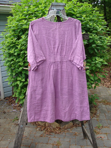Barclay Linen Posey Dress in Tiny Floral Orchid, size 0, hangs on a wooden frame. Features include elbow-length sleeves, oval neckline, flowy shape, exterior pockets, and rear elastic tab.