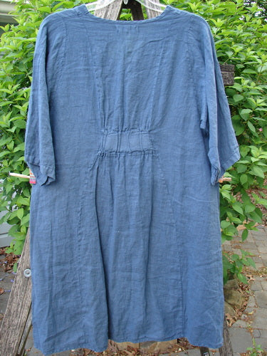 Barclay Linen Posey Dress Daisy Ray Blue Echo Size 0 displayed on a wooden stand, featuring elbow-length sleeves, an oval neckline, flowy figure-eight shape, and two drop front pockets amidst greenery.