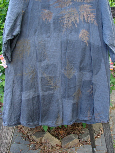 Barclay Linen Lucida Top Metallic Fern Blue Storm Size 0, a flowy, steel-blue tunic with brown fern imprints, hangs on a wooden rod outdoors, showcasing its lightweight linen and artistic design.