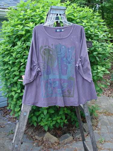 Barclay Draw Gather Sleeve Cafe Top Power Plum Size 0, featuring a stylized cactus graphic, gathered sleeves, and rolled edges, displayed outdoors on a wooden ladder against a leafy backdrop.