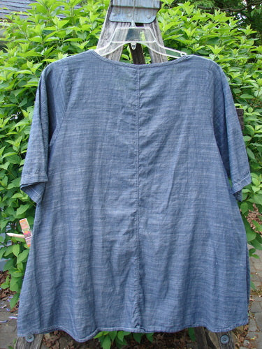 Barclay Chambray Short Sleeved Top featuring a floral joy theme in dark denim striation, displayed on a hanger outdoors. The tunic has a graduated A-line shape and rounded hemline.