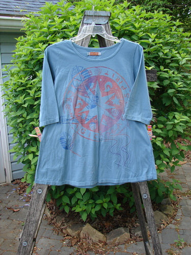 Barclay Three Quarter Sleeved Tee Metallic Belief Spirit Magic Sky Blue Size 1 draped over a wooden ladder outdoors, showcasing its circular graphic and rolled neckline against a natural backdrop.