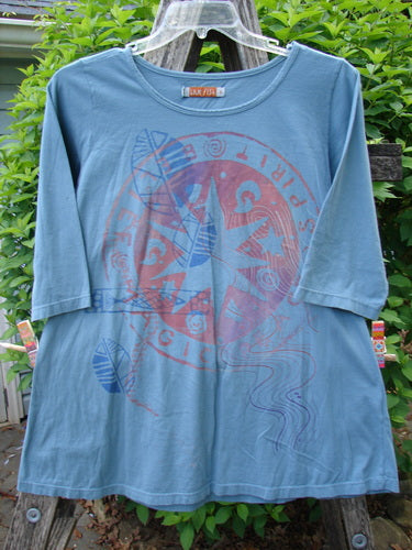 Barclay Three Quarter Sleeved Tee Metallic Belief Spirit Magic Sky Blue Size 1 features a circular graphic with arrows and leaf motifs, hanging outdoors on a clear hanger against greenery.