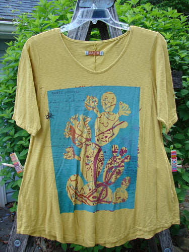 Barclay Slub Short Sleeved Travel Tee Cactus Garden Citron Size 1 features a cactus graphic in teal on a mustard-yellow backdrop, showcased hanging outdoors against green foliage.
