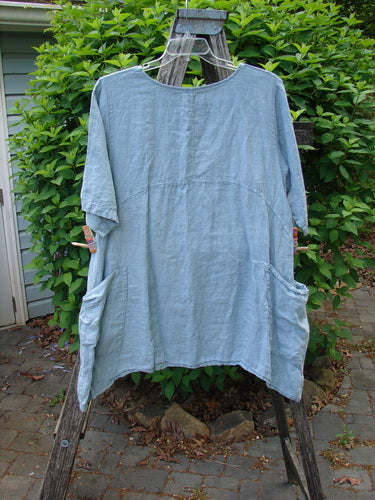 Barclay Linen Urchin Dress Leaf Floral Dusky Watercolor Size 1 displayed on a wooden ladder, featuring elbow-length sleeves, large hip-level pockets, and a downward curved empire waist seam with varying side lengths.
