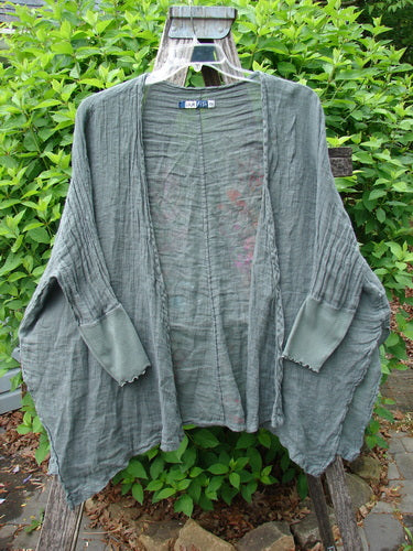 Barclay Crinkle Linen Open Front Rib Sleeve Cheerful Cardigan in Caper, featuring dolman sleeves, ribbed cuffs, and a scripted fern design, displayed on a wooden hanger against leafy shrubs.