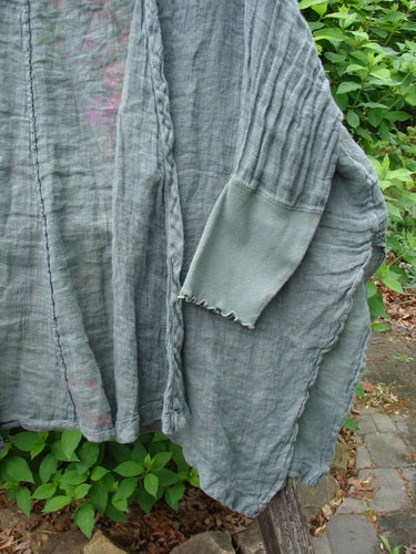 Barclay Crinkle Linen Open Front Rib Sleeve Cheerful Cardigan Scripted Fern Caper Size 0, featuring dolman sleeves, ribbed cuffs, pintuck details, and raw-edge hems, drapes over a wooden bench outdoors.