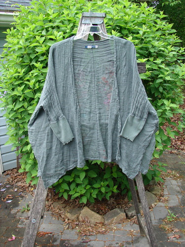 Barclay Crinkle Linen Open Front Rib Sleeve Cheerful Cardigan Scripted Fern Caper Size 0 displayed draped over a wooden trellis, showcasing dolman sleeves and a vented hemline against a leafy shrub backdrop.