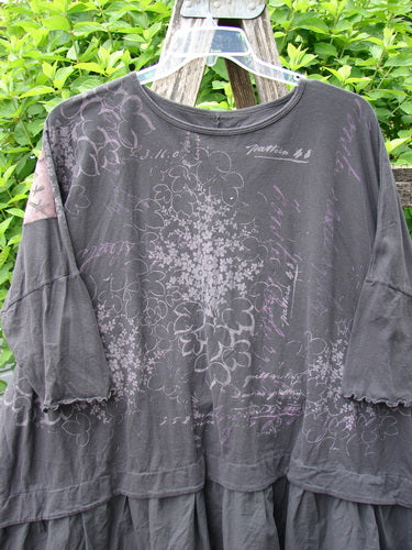 Barclay Batiste Flounce Esperanza Tunic in Storm, showcasing dolman sleeves with lettuce edges, layered hem, and faded floral script print, draped over a ladder.
