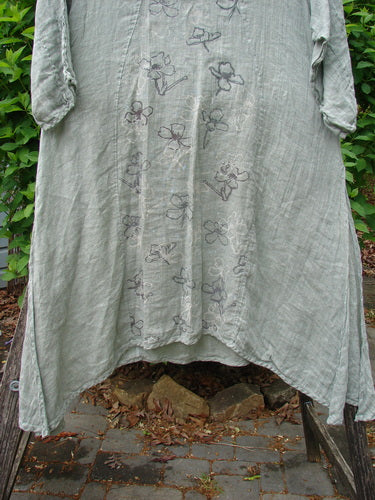Barclay Linen High Vent Venetian Dress Contour Floral Sage Size 1, featuring embroidered floral motifs, three-quarter sleeves, oval neckline, and varying hemline, hangs on a rustic frame against leafy greenery.