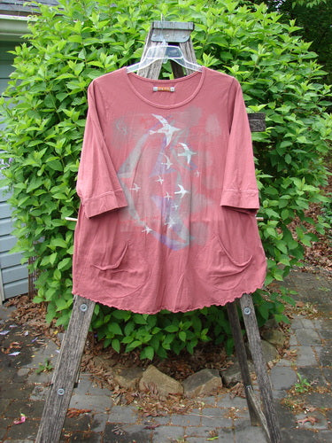 Daisy Hagey Barclay Banded Artist Pocket Top Metallic Bird Flight Rust Size 1 in rust-toned fabric, with a metallic bird graphic, short sleeves, and front pockets, hanging on a wooden stand outdoors.