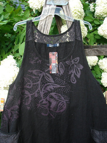 Bluefishfinder.com Barclay NWT Summercloth Lace Two Pocket Pinafore Floral Fern Black Size 0, a black sleeveless pinafore with lace-trimmed straps and a sheer floral fern overlay. The garment hangs on a clear hanger against dense green leaves and white blossoms, with a soft matte-black body and a visible tag at the chest.