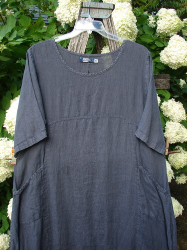 Bluefishfinder.com Barclay Linen Draw Bottom Urchin Dress in unpainted dusty navy, size 0, shown as a loose linen dress with a rounded neckline, short sleeves, and gathered side panels that create a soft, draped silhouette. The dark blue fabric hangs against a leafy floral backdrop.