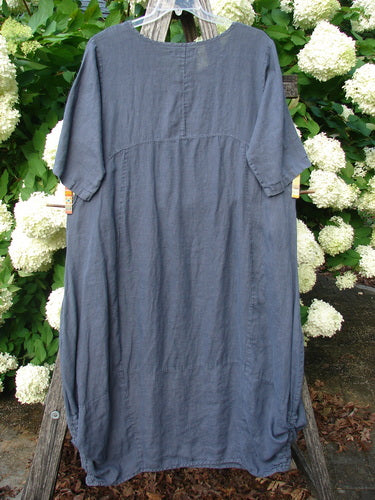 Bluefishfinder.com Barclay Linen Draw Bottom Urchin Dress in unpainted dusty navy, a loose short-sleeve linen dress with a softly gathered waist, curved seam detailing, and a drawstring hem. It hangs front-facing against leafy white hydrangeas, with a muted, textured finish.