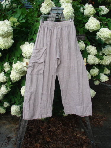 Bluefishfinder.com Barclay Linen Single Side Pocket Banded Bottom Pant in unpainted mallow, size 0, shown as loose, cropped linen pants with a banded waist and a single side pocket. The pale taupe fabric hangs from a rustic wooden ladder before white hydrangeas, creating a soft, natural contrast.