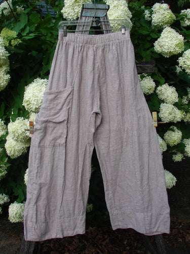Bluefishfinder.com Barclay Linen Single Side Pocket Banded Bottom Pant in unpainted mallow, size 0, shown as loose linen pants with a single side pocket, banded hems, and a softly wrinkled matte texture. The muted taupe fabric hangs straight on a hanger against dense white hydrangea blooms and dark green foliage.