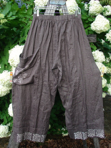 Susan Crawford Barclay linen single crop side pocket banded bottom pant in mocha, with Celtic floral trim and size 2, hanging on a hanger against green foliage and white flowers.