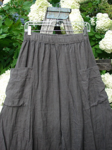Bluefishfinder.com Barclay Linen Tie Bottom 4 Square Pant Cross Dye Army Unpainted Size 0, shown as dark gray linen pants with a tie waist, four square patch pockets, and a softly crinkled texture. The garment hangs from a clear plastic hanger against leafy white flowers.