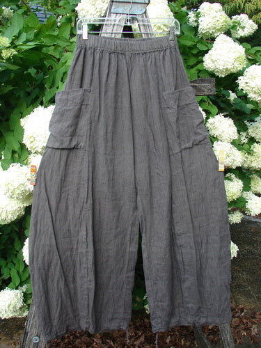 Bluefishfinder.com Barclay Linen Tie Bottom 4 Square Pant Cross Dye Army Unpainted Size 0 in washed gray linen, with a loose wide-leg silhouette, gathered waistband, and large square cargo pockets. The pants hang from a hanger against dense green foliage and white blooms.