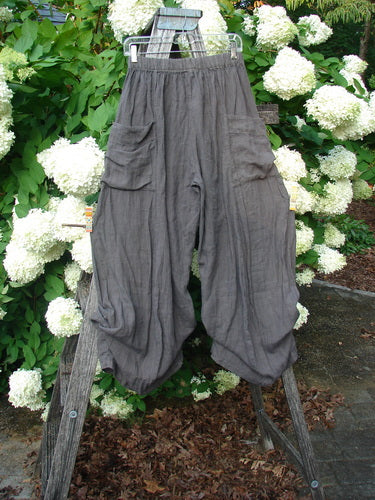 Bluefishfinder.com Barclay Linen Tie Bottom 4 Square Pant Cross Dye Army Unpainted Size 0 in cross-dyed gray linen, with an elastic waist, loose balloon legs, and tie hems. The pants hang on a wooden ladder amid white hydrangeas, with soft daylight and a natural green backdrop.