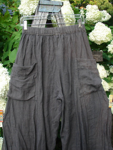 Bluefishfinder.com Barclay Linen Tie Bottom 4 Square Pant in cross-dye army, unpainted size 0, with a loose, crinkled linen texture and wide legs. The dark gray pants hang from a clear hanger, showing oversized square patch pockets against leafy white hydrangeas.