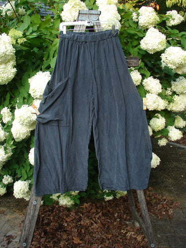 Bluefishfinder.com Barclay Silk Dimensional Pant in unpainted black, size 1, with a loose, wide-leg silhouette, gathered waistband, and large side pocket. The textured silk drapes over a wooden hanger, set against dense green foliage and white hydrangea blooms.