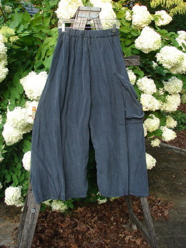 Bluefishfinder.com Barclay Silk Dimensional Pant in unpainted black, size 1, with a loose drape, gathered elastic waist, and a side cargo pocket. The dark silk fabric hangs from a wooden ladder outdoors, set against dense green leaves and white hydrangea blooms.