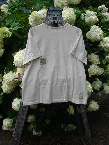 Bluefishfinder.com 1999 Short Sleeved Tee Flower Tops Natural Size 1 in a soft natural beige, with a loose short-sleeve silhouette, crew neckline, and subtle printed flower details on the chest and hem. The tee hangs from a weathered wooden stand before dense white hydrangea blooms and dark green foliage, creating a layered, outdoor display.