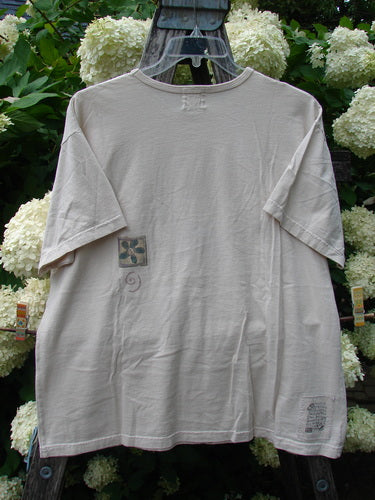 Bluefishfinder.com 1999 Short Sleeved Tee Flower Tops Natural Size 1, a natural-toned short-sleeve T-shirt with a loose, boxy drape and soft, lightly wrinkled cotton texture. It hangs centered on a dark hanger amid white hydrangeas, with small printed graphics on the chest and hem.