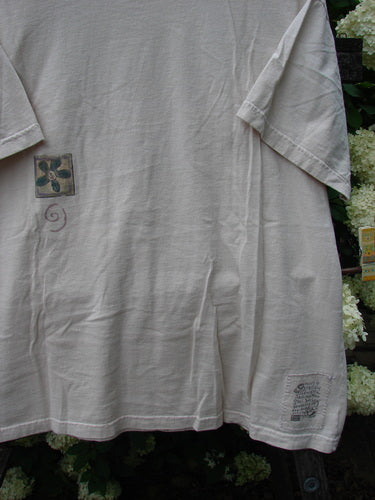 Bluefishfinder.com 1999 Short Sleeved Tee Flower Tops Natural Size 1 in soft natural beige cotton, shown front-on with short sleeves, a loose drape, a small flower graphic on the chest, and a tiny printed mark near the hem.
