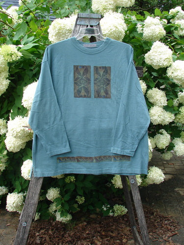 Bluefishfinder.com 1997 Long Sleeved Tee Double Celtic Flower Cypress Size 2 in muted cypress blue, a long-sleeve T-shirt with a soft matte finish, centered double Celtic flower graphic, and a narrow patterned hem band. It hangs front-facing on a wooden ladder amid white hydrangeas and dense green foliage.